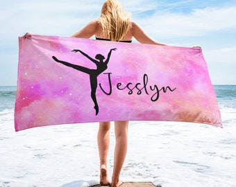 Könnte beinhalten: Ein rosa Strandtuch mit Aquarell-Effekt, das eine schwarze Silhouette einer Ballerina in anmutiger Pose zeigt. Der Name "Jesslyn" ist in Schreibschrift geschrieben. Das Handtuch wird an einem Strand mit dem Meer im Hintergrund gehalten.