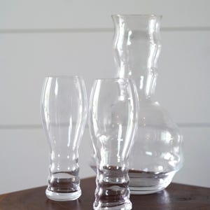 May include: Three clear glass containers, two are tall and narrow with a textured base, and one is a wider vase with a rounded base. The containers are on a wooden surface.