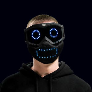 May include: A person wearing a black hoodie and a futuristic black mask with blue LED lights. The mask has a smiling face design with lights for eyes and a mouth. The person is against a dark blue background.
