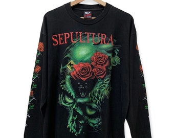Puede incluir: Camiseta negra de manga larga con un gráfico de calavera verde y rosas rojas. El texto "SEPULTURA" está impreso en rojo en la parte delantera de la camiseta.