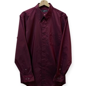 May include: A long-sleeved, burgundy button-down shirt with a single chest pocket. The shirt has a classic collar and is hanging on a wooden hanger.