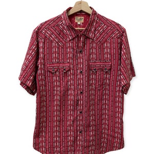 Puede incluir: Una camisa de manga corta abotonada a rayas rojas y blancas con cuello de estilo western y dos bolsillos de solapa. La camisa tiene una pequeña etiqueta que dice "Two Palms".