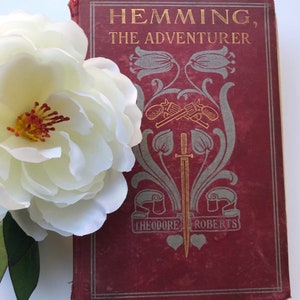 May include: A vintage hardback book titled "Hemming, The Adventurer" by Theodore Roberts. The book cover is a deep red with gold lettering and an intricate design featuring crossed swords, a dagger, and stylised flowers.
