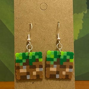 May include: A pair of green and brown Minecraft-themed earrings. The earrings are made of perler beads and feature a block of dirt with grass on top.