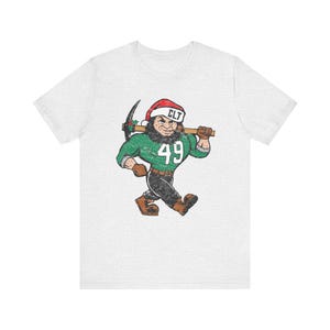 May include: A light grey short-sleeve t-shirt featuring a graphic of a bearded character wearing a Santa hat with the text CLT, a green jersey with the number 49, and black pants. The character carries a pickaxe over their shoulder.