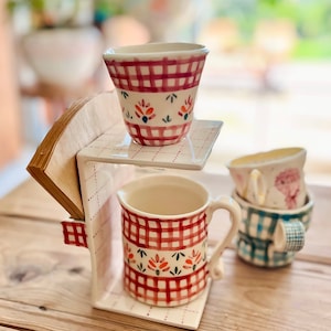 Op de afbeelding: Een keramische bekerhouder met een rood-wit geruit patroon. De houder heeft een plank voor een koffiefilter en ruimte voor een beker. Er staan twee bekers op de houder, een met een rood-wit geruit patroon en de andere met een blauw-wit geruit patroon.