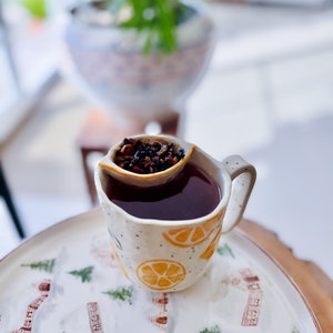 Puede incluir: Una taza de cerámica blanca con un diseño de limón y un infusor integrado lleno de hojas de té sueltas. La taza está sobre un plato blanco con un diseño de escena de invierno.