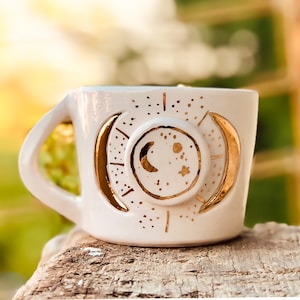 Op de afbeelding: Witte keramische mok met een gouden halve maan en sterrenontwerp. De mok heeft een handvat en is perfect voor koffie of thee.
