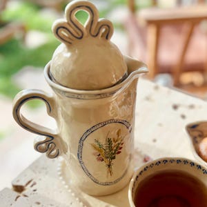 May include: A cream-coloured ceramic teapot with a lid and handle, decorated with blue trim and a floral design. The teapot sits on a matching tray, with a teacup filled with tea. The design is reminiscent of vintage tableware.