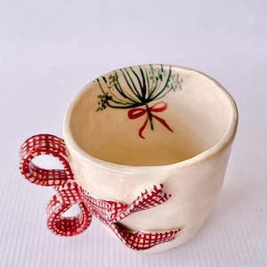 May include: A white ceramic mug with a red and white gingham bow handle. The mug has a green and red floral design inside.