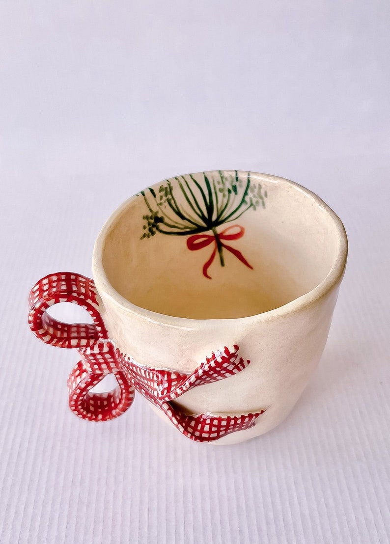 May include: A white ceramic mug with a red and white gingham bow handle. The mug has a green and red floral design inside.