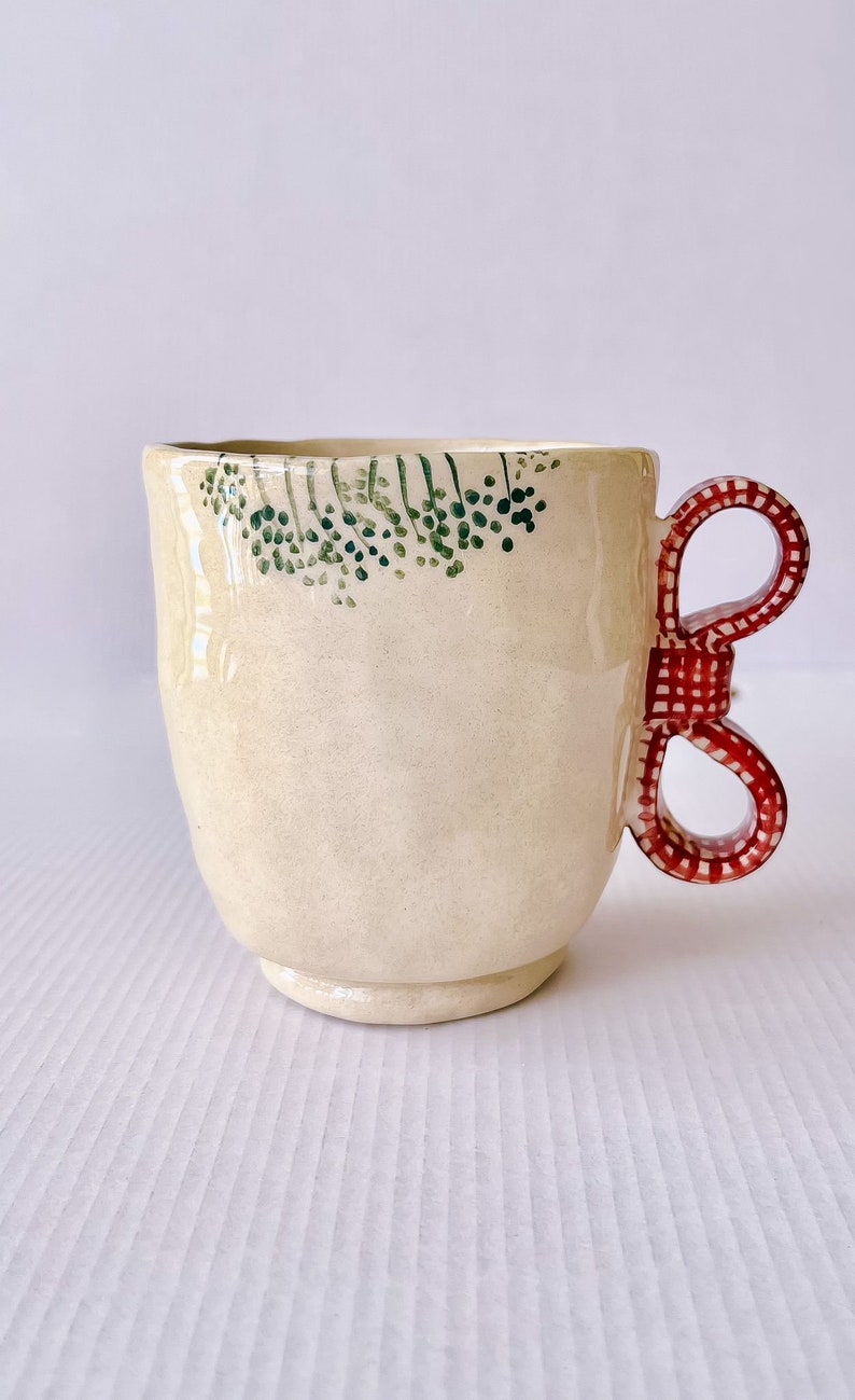 May include: A cream-colored ceramic mug with a green floral design around the rim and a red and white bow-shaped handle.