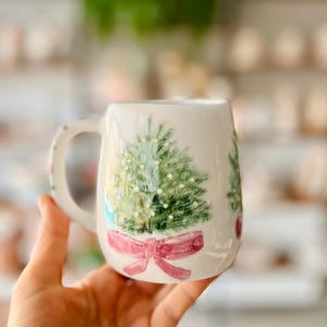 Puede incluir: Taza de cerámica blanca con un diseño pintado a mano de un árbol de Navidad verde con un lazo rosa. La taza tiene un asa y es perfecta para bebidas calientes.