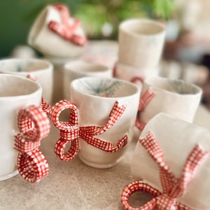 May include: A collection of white ceramic mugs with red and white gingham ribbon bows. The mugs are arranged in a group on a white surface.
