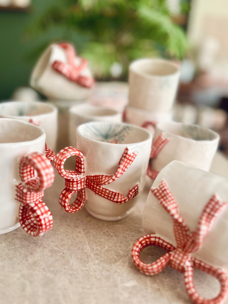 May include: A collection of white ceramic mugs with red and white gingham ribbon bows. The mugs are arranged in a group on a white surface.