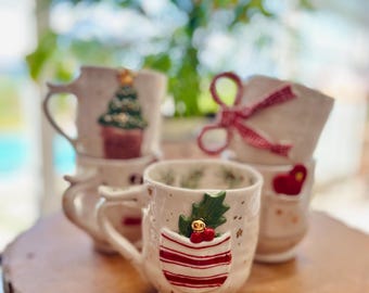May include: Set of four white ceramic mugs with festive Christmas designs. Each mug features a different holiday-themed decoration, including a Christmas tree, a bow, a stocking, and a holly sprig. The mugs are arranged on a wooden surface.