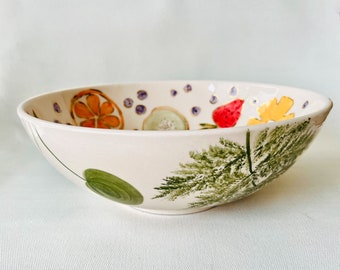 May include: A white ceramic bowl with a green fern design and painted fruit and flowers. The bowl is decorated with orange slices, a strawberry, and a yellow flower.