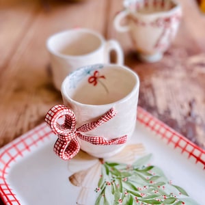 May include: A white ceramic teacup with a red and white checkered bow on the handle. The cup is sitting on a white tray with a red and white checkered border and a green and red painted holly sprig.