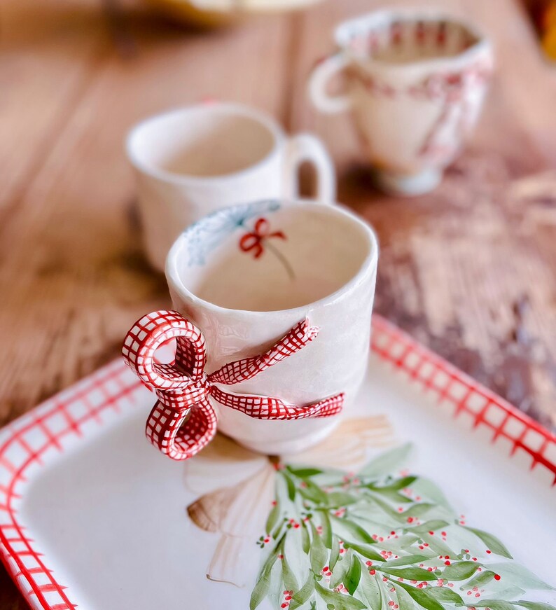 May include: A white ceramic teacup with a red and white checkered bow on the handle. The cup is sitting on a white tray with a red and white checkered border and a green and red painted holly sprig.