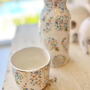 Puede incluir: Un juego de jarrón y taza de cerámica blanca. Ambas piezas están adornadas con un delicado estampado floral en tonos azules, naranjas y verdes. Los detalles dorados gotean por los bordes, añadiendo un toque de elegancia. El jarrón es más alto que la taza.