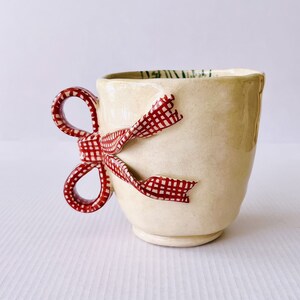 May include: A ceramic mug with a red and white gingham bow handle. The mug is cream colored and has a textured surface.