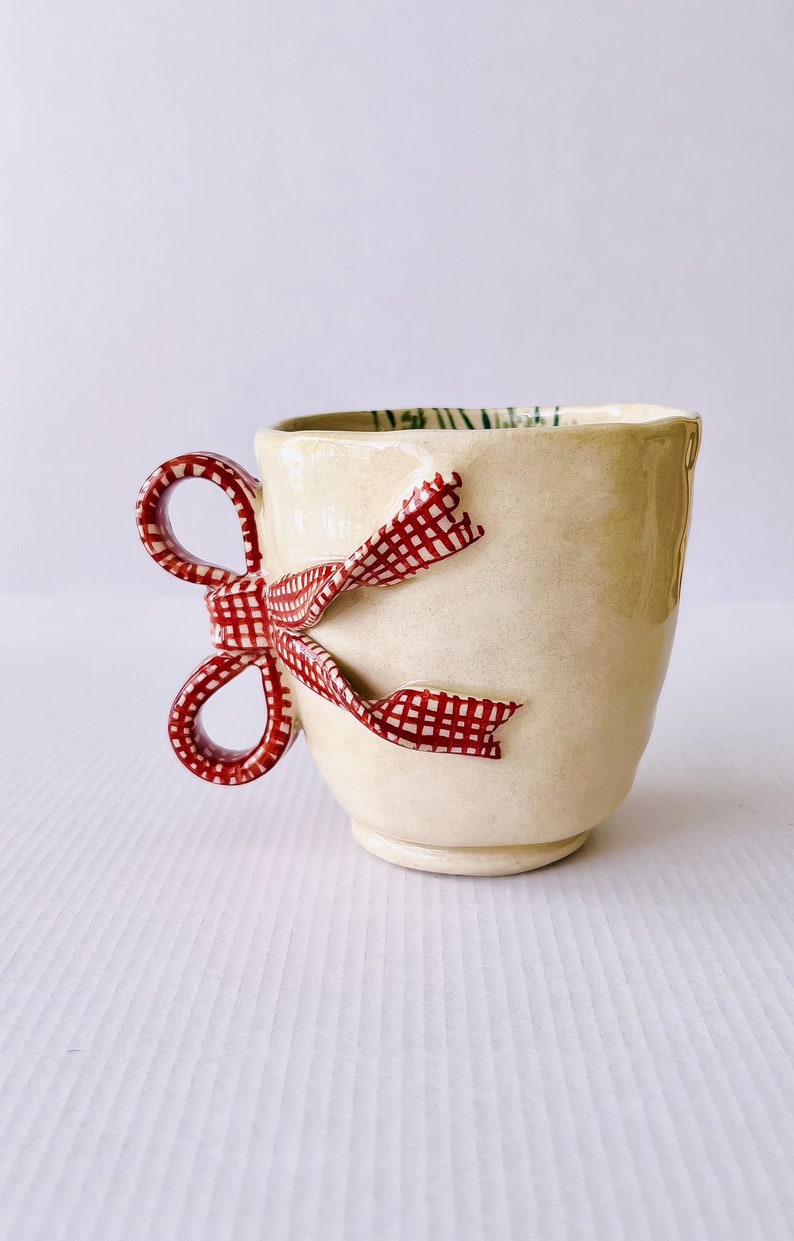 May include: A ceramic mug with a red and white gingham bow handle. The mug is cream colored and has a textured surface.