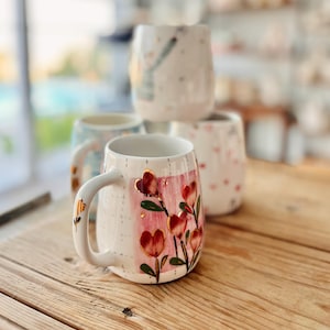 Puede incluir: Una taza de cerámica blanca con un diseño floral rosa y dorado con corazones rojos. La taza tiene un asa blanca y está sobre una superficie de madera.