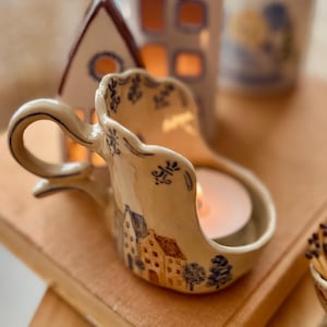 Könnte beinhalten: Cremefarbener Keramik-Kerzenhalter mit Griff, verziert mit einem Dorfmotiv. Enthält eine brennende Teelichtkerze. Weitere Keramikhäuser im Hintergrund bilden eine gemütliche Dekoration. Der Halter ist ca. 15 cm lang.