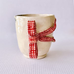 May include: A white ceramic mug with a red and white gingham ribbon handle. The ribbon is tied in a bow and the mug is decorated with a floral pattern.
