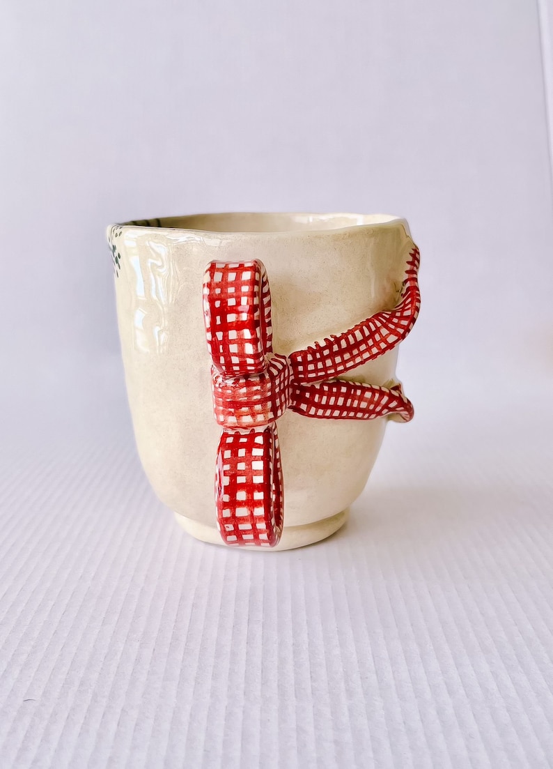 May include: A white ceramic mug with a red and white gingham ribbon handle. The ribbon is tied in a bow and the mug is decorated with a floral pattern.