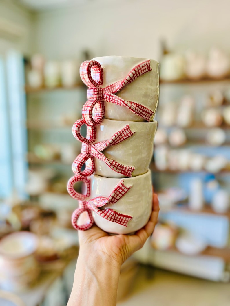 May include: Stack of four white ceramic mugs with red and white gingham ribbon bows. The mugs are stacked in a hand.