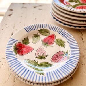 May include: White ceramic plates with a hand-painted strawberry design. Each plate has a blue striped border and illustrations of ripe strawberries and green leaves. A stack of similar plates is in the background.