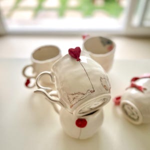 Może przedstawiać: Kolekcja białych ceramicznych kubków i mały imbryk. Każdy kubek ma uchwyt w kształcie serca i czerwony akcent w kształcie serca. Jeden kubek ma wzór czerwonego balonu w kształcie serca. Imbryk ma czerwony akcent w kształcie serca.