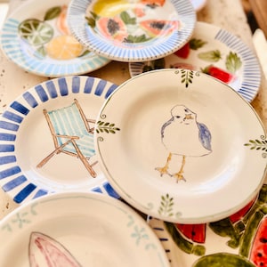 May include: A collection of hand-painted ceramic plates with various designs. Some plates feature fruit illustrations like watermelon, papaya, and citrus. Others showcase a beach chair and a seagull. The plates have blue striped borders and decorative edges.