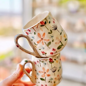 Puede incluir: Dos tazas de cerámica blanca con un patrón floral y detalles dorados. Las tazas están apiladas una encima de la otra. El patrón floral incluye flores rosas, rojas, azules y verdes.