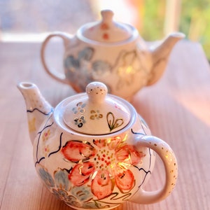 Op de afbeelding: Twee witte theepotten van keramiek met bloemmotieven. De theepot op de voorgrond heeft een roze en gouden bloemenmotief, terwijl de theepot op de achtergrond een blauw en gouden bloemenmotief heeft.