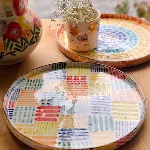 Puede incluir: Dos platos de cerámica con coloridos patrones geométricos. El plato más grande tiene un diseño de patchwork con líneas y puntos rojos, amarillos, azules y verdes. El plato más pequeño tiene un diseño circular con un arcoíris de colores.