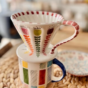 Op de afbeelding: Een witte keramische koffiekan met een rood, wit en groen bloemenpatroon staat op een witte keramische mok met een blauwe handgreep en een kleurrijk geometrisch patroon. De mok en de kan staan op een geweven rieten oppervlak.