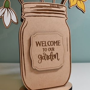 Może przedstawiać: Drewniana dekoracja w kształcie słoika typu Mason z napisem &bdquo;WELCOME TO OUR garden&rdquo; na etykiecie. Słoik zawiera drewniane wycięcia kwiat&oacute;w w kolorze ż&oacute;łtym, białym i r&oacute;żowym. Podstawa to okrągła, drewniana podstawka z czarną krawędzią.