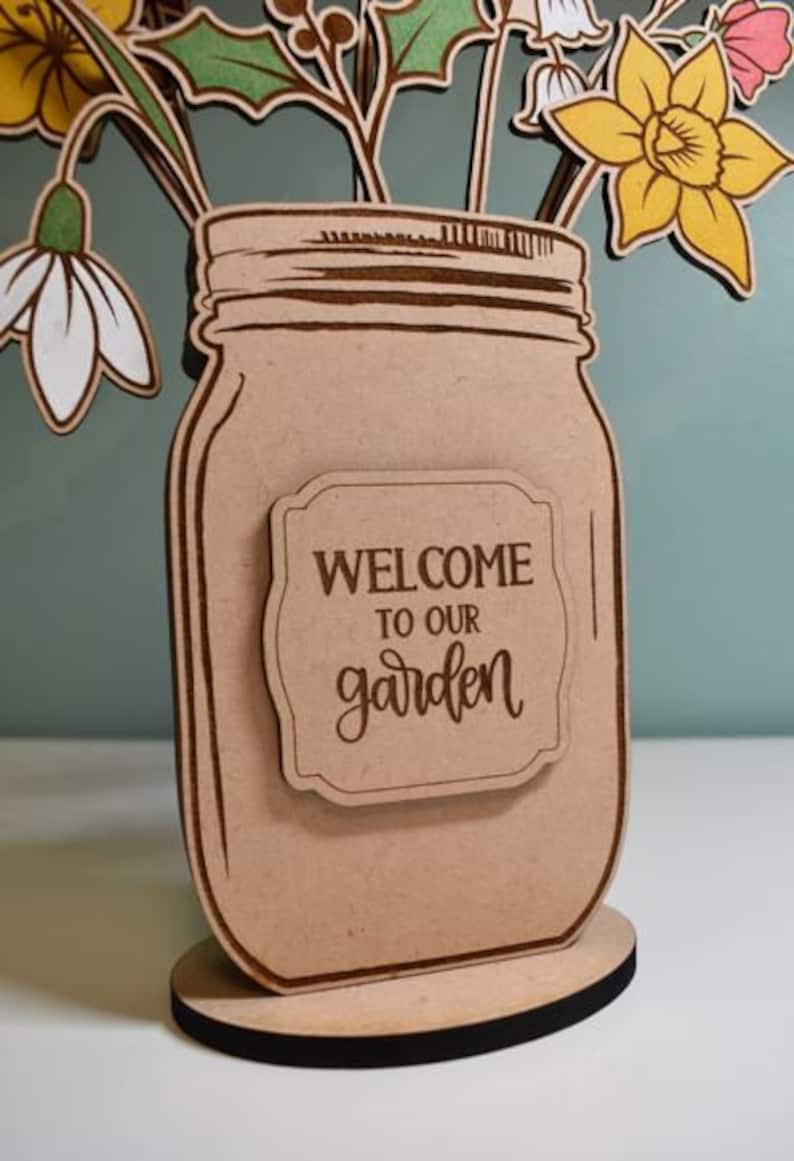 Może przedstawiać: Drewniana dekoracja w kształcie słoika typu Mason z napisem &bdquo;WELCOME TO OUR garden&rdquo; na etykiecie. Słoik zawiera drewniane wycięcia kwiat&oacute;w w kolorze ż&oacute;łtym, białym i r&oacute;żowym. Podstawa to okrągła, drewniana podstawka z czarną krawędzią.