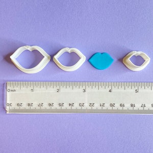Pode incluir: Três cortadores de biscoitos de plástico branco em forma de lábios, em tamanhos diferentes, e um recorte de fondant azul em forma de lábios. Os cortadores estão sobre uma superfície roxa com uma régua.