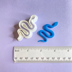 May include: Two snake-shaped cookie cutters, one white and one blue, are shown on a purple surface with a ruler for scale. The white cutter is larger and has a more intricate design. The blue cutter is smaller and has a simpler design.