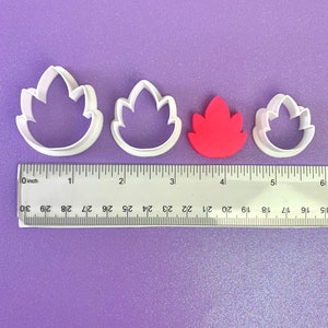 May include: Three white plastic cookie cutters shaped like leaves, with a red fondant leaf in the center. The cutters are arranged in a row from largest to smallest. A ruler is visible below the cutters.