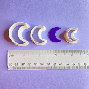 May include: Four crescent moon cookie cutters, two white plastic and two purple plastic. The cutters are arranged in a row from largest to smallest. A ruler is placed below the cutters to show their size.