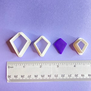 May include: Four white plastic diamond-shaped cookie cutters of different sizes, with a purple diamond-shaped piece of fondant in the middle, on a purple surface with a ruler.
