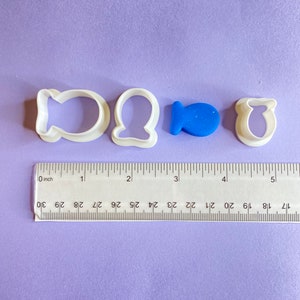 May include: Four white plastic cookie cutters in the shape of a fish, a seashell, a whale, and a starfish. A blue fish-shaped cookie cutter is also included. The cookie cutters are on a purple surface with a ruler for scale.