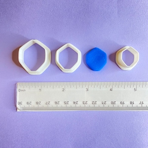 May include: Four white plastic geometric shape cookie cutters and a blue geometric shape are arranged on a purple surface next to a ruler. The largest cutter is approximately 7.6 cm across.