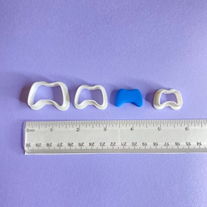 May include: Three white plastic cookie cutters shaped like a video game controller, with the largest cutter measuring 2 inches wide. A smaller blue plastic cookie cutter is also included, measuring 1 inch wide.