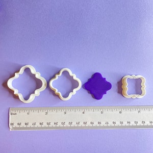 Pode incluir: Quatro cortadores de biscoitos em tamanhos diferentes, dois são brancos e dois são roxos. Os cortadores têm a forma de um quadrado com cantos arredondados e um design decorativo. Uma régua está na parte inferior da imagem.
