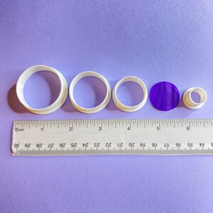 Puede incluir: Cuatro cortadores de galletas de plástico blanco en diferentes tamaños, un círculo morado y un tubo de plástico blanco, todo sobre una superficie morada con una regla que mide 15,24 cm.